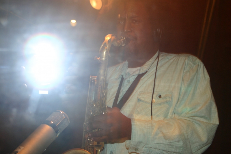 Kamasi Washington in a white long sleeved shirt, playing the saxophone on a live stage, with a bright light shining behind him.
