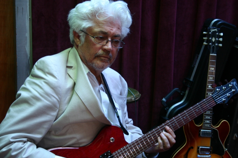 Larry Coryell in a white coat with his eyes closed, playing the guitar. 